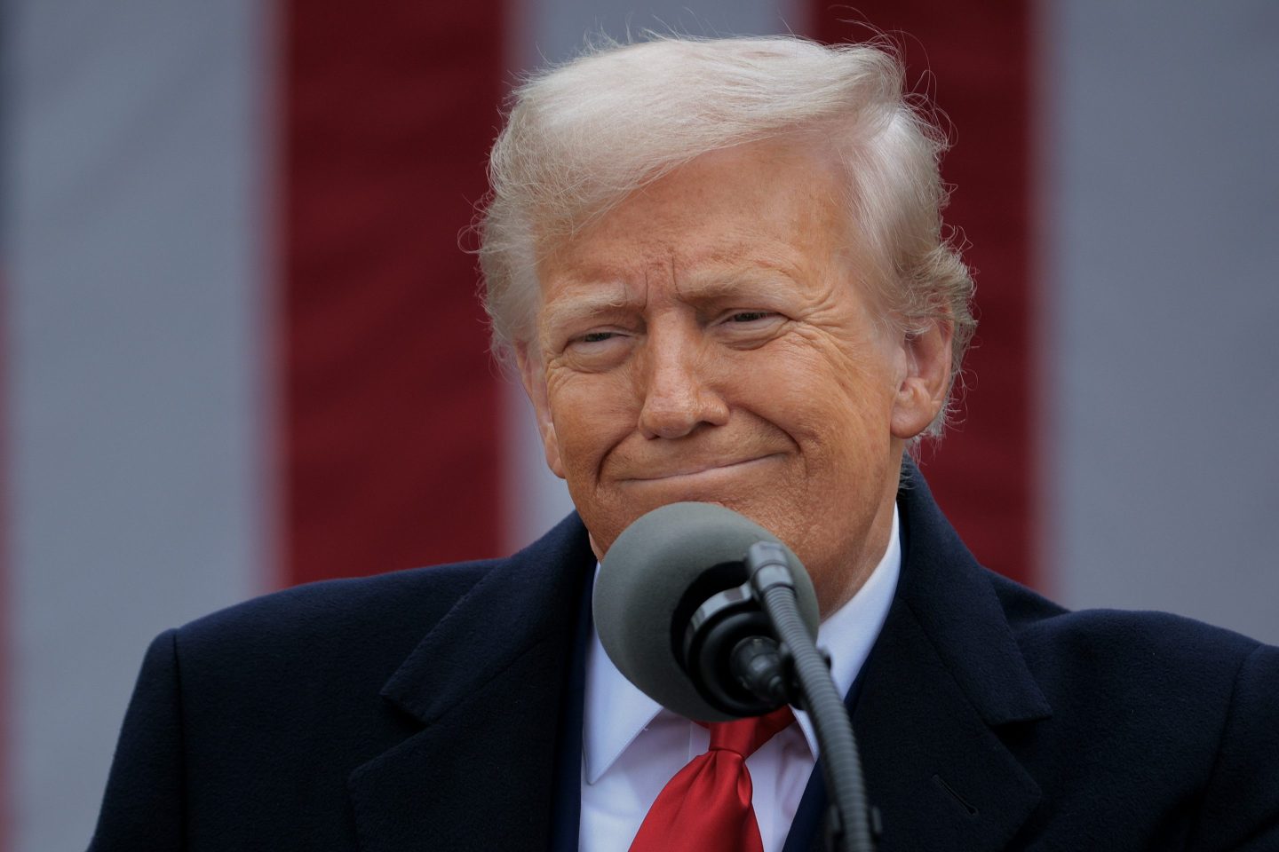 President Donald Trump smirks just off the camera in a black suit with a red tie.