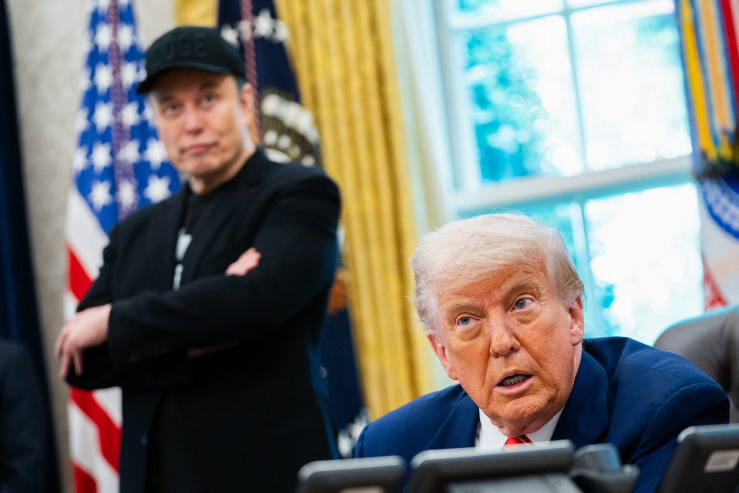 Elon Musk, in a black sports jacket and black hat, stands behind Trump and looks over crossed-arms as Trump speaks to reporters.