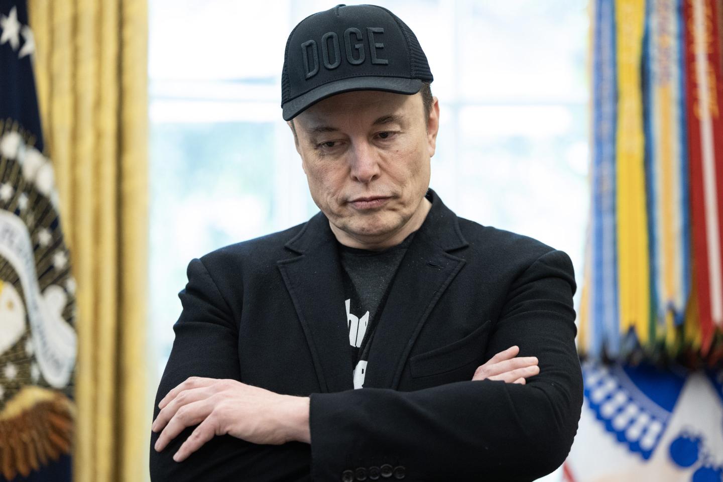 Elon Musk, sporting a black sports jacket and a black 'DOGE' hat, looks down during a news conference with President Donald Trump on May 30, 2025 inside the Oval Office at the White House in Washington.