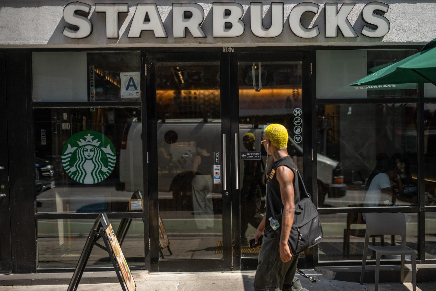 A young person with yellow hair and wired headphones walks in front of the Starbucks coffee shop, looking in.
