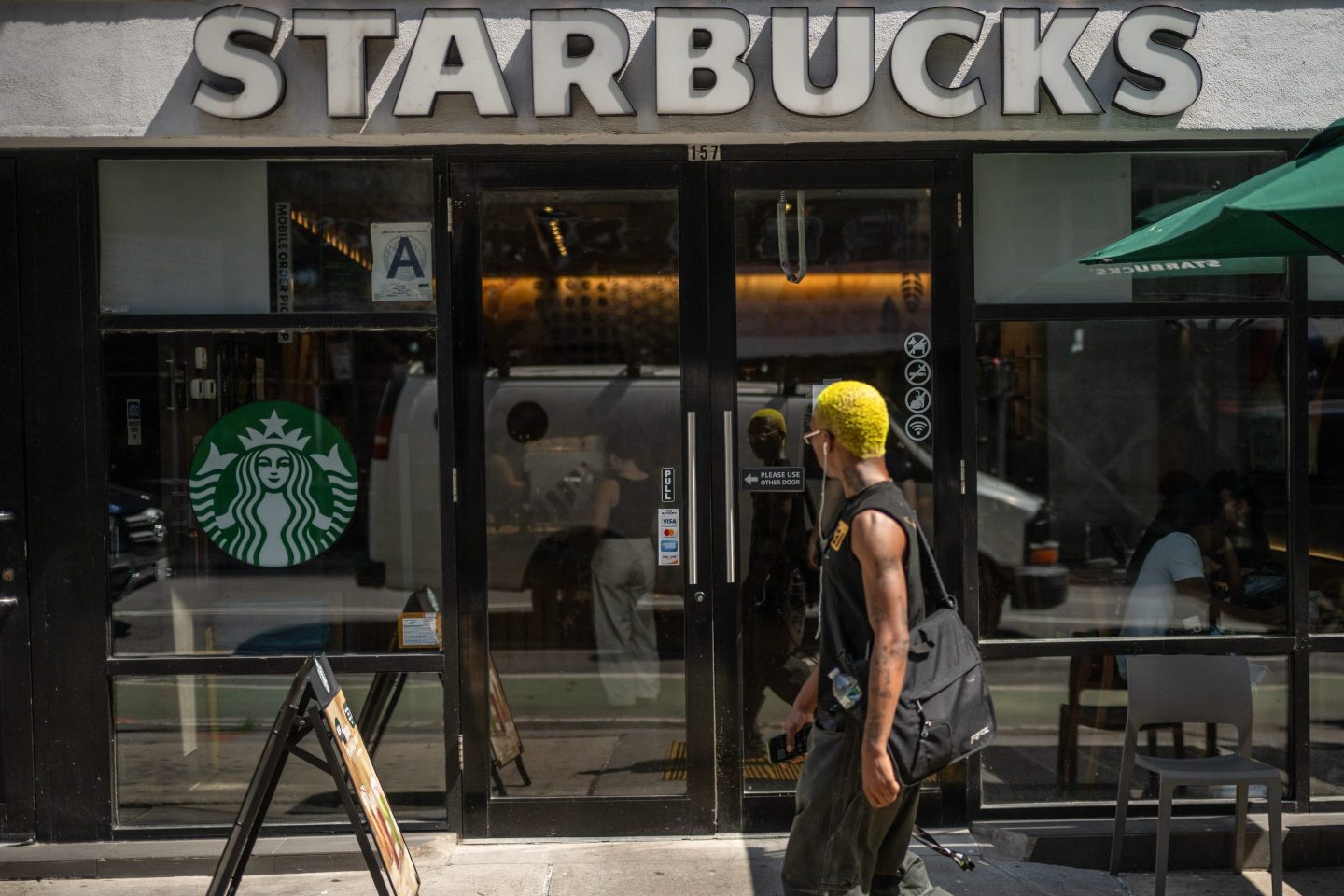 A young person with yellow hair and wired headphones walks in front of the Starbucks coffee shop, looking in.