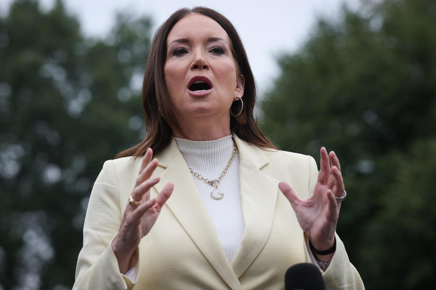 Brooke Rollins, standing outside, speaks and gestures with both hands in front of her.