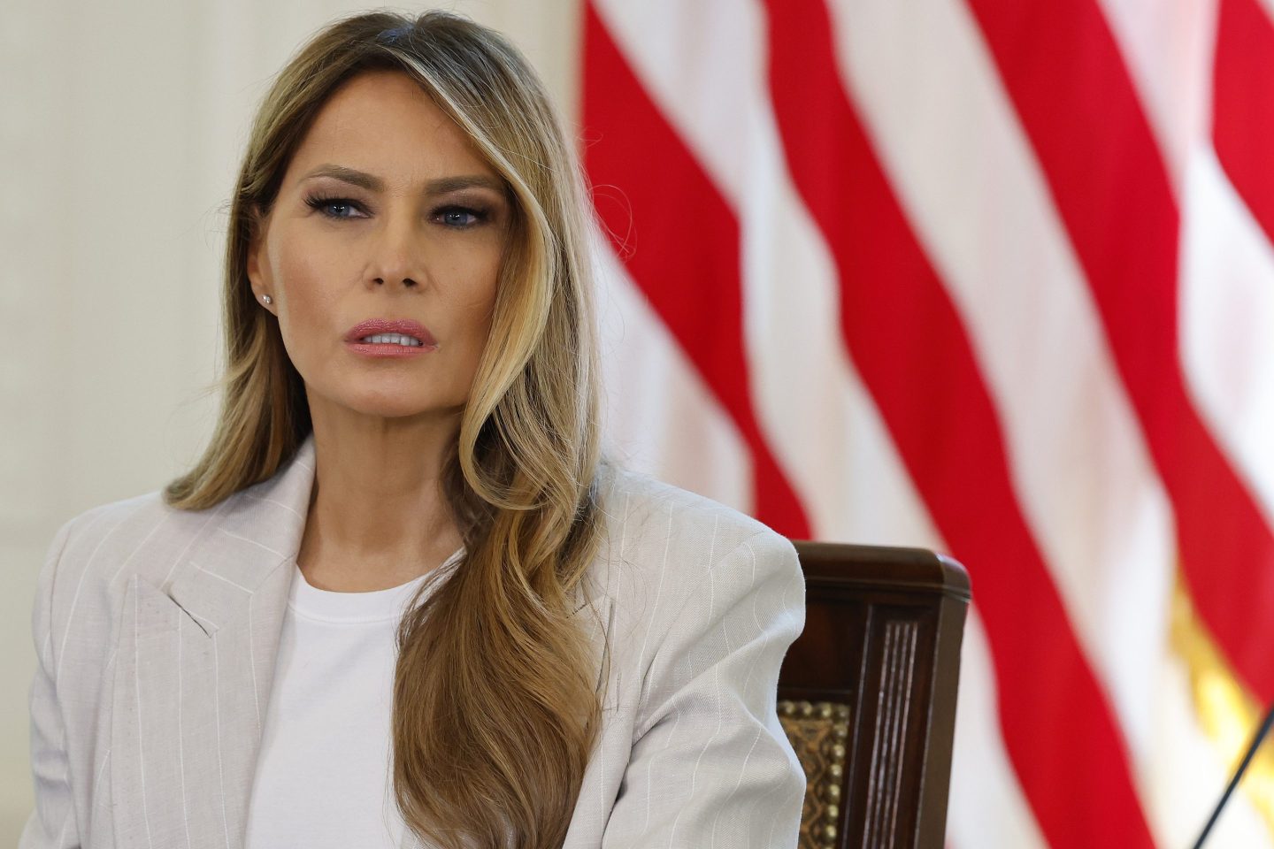 A woman in a light gray suit jacket and white t-shirt sits in front of an American flag