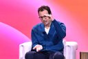 Dario Amodei, sitting in a white chair in front of a pink background on stage, puts both hands out in front of him, gesturing.