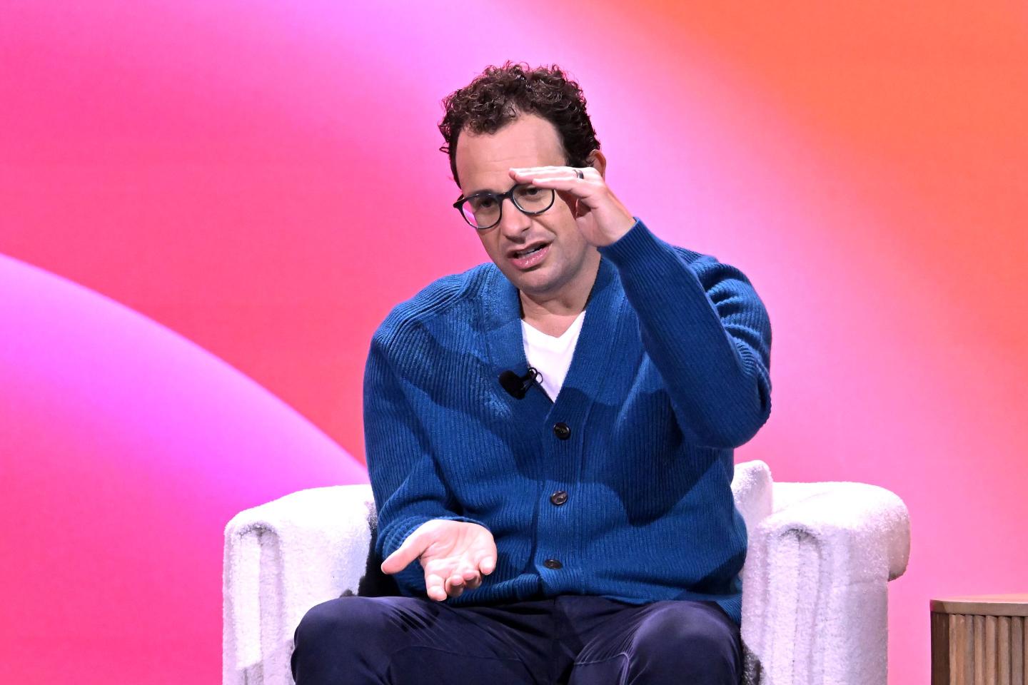 Dario Amodei, sitting in a white chair in front of a pink background on stage, puts both hands out in front of him, gesturing.