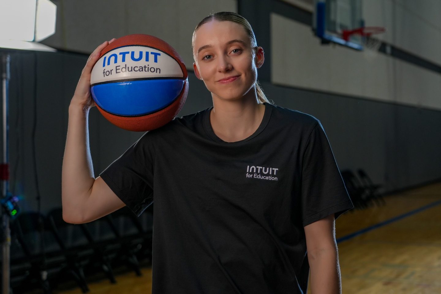 Paige Bueckers holds a blue and white basketball on her shoulder that reads, "Intuit for education."