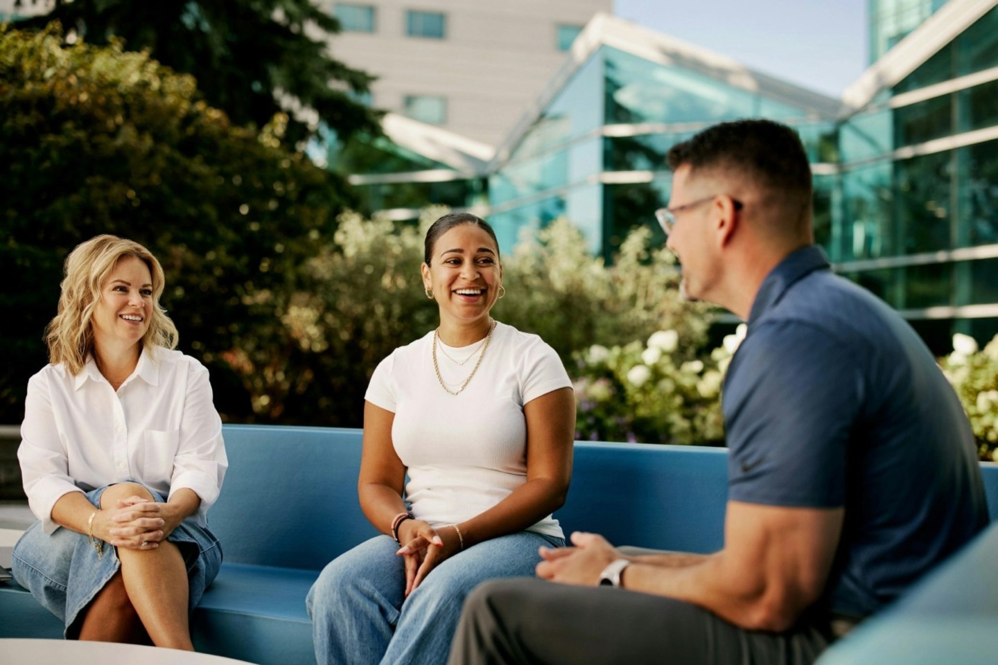 Three people sit on a couch smiling and talking with each other.