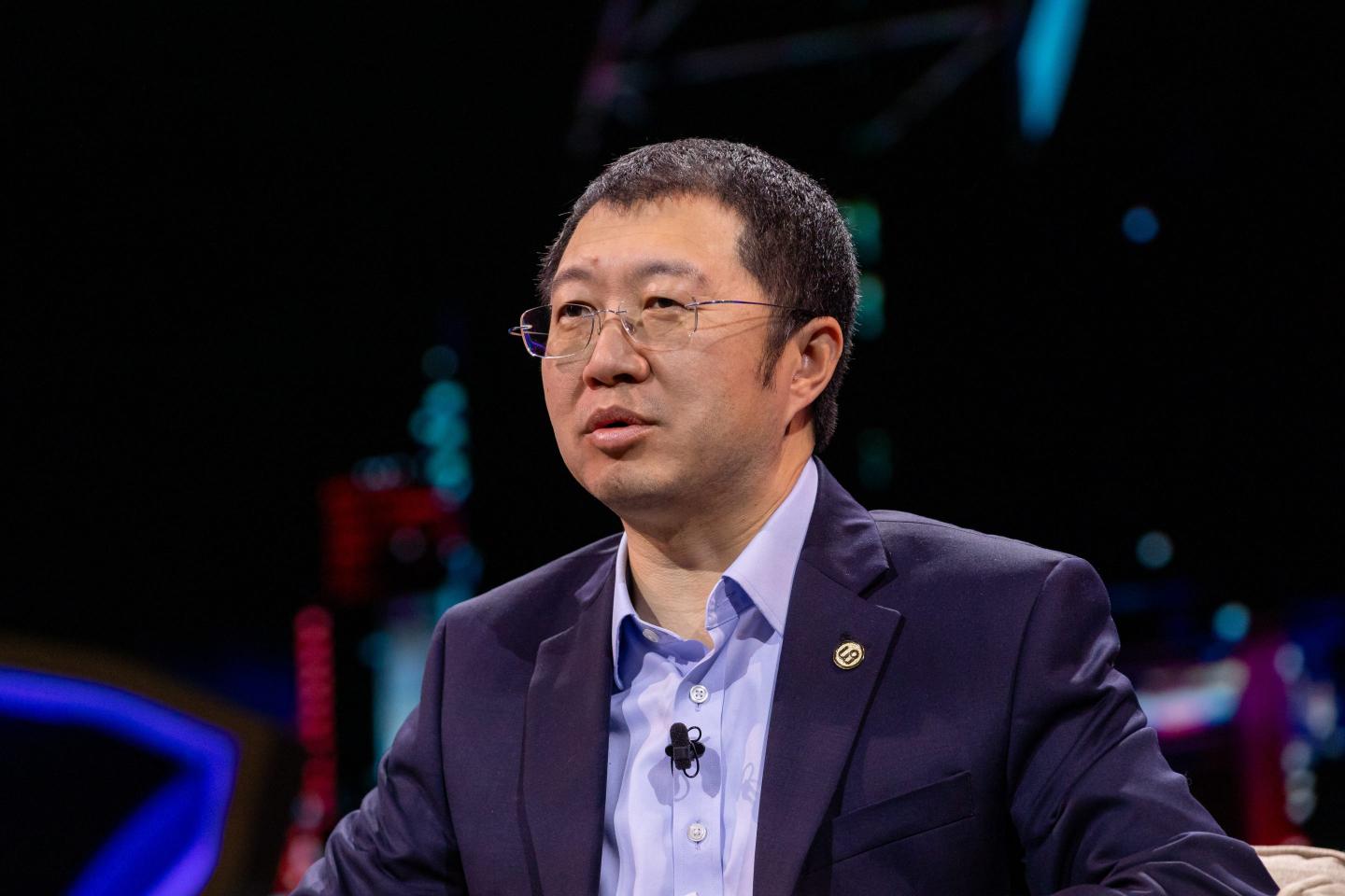 Ton Han, wearing a dark suit and purple shirt, speaks in front of a dark background.