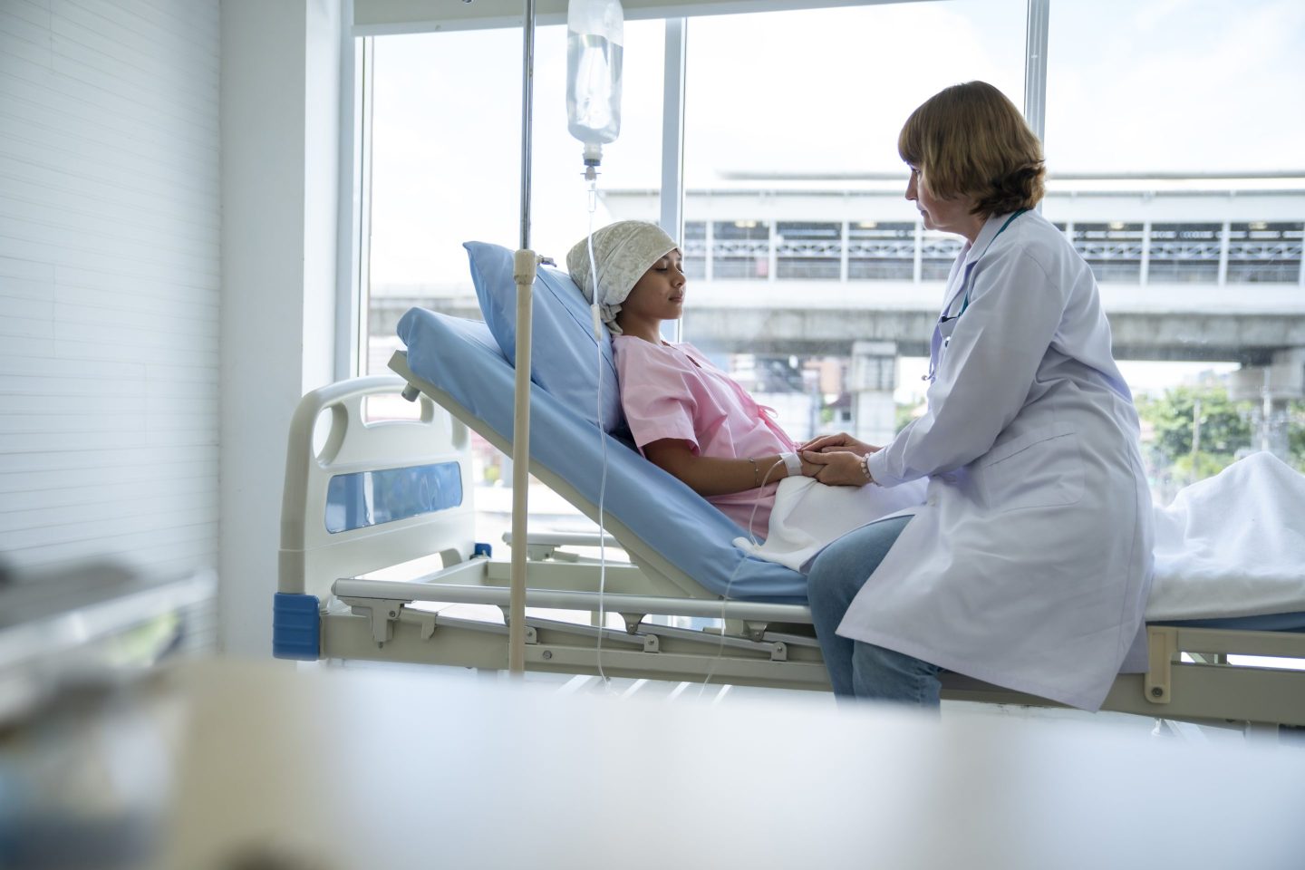 A female doctor encourages a young woman with cancer by holding hands on the patient's bed.
