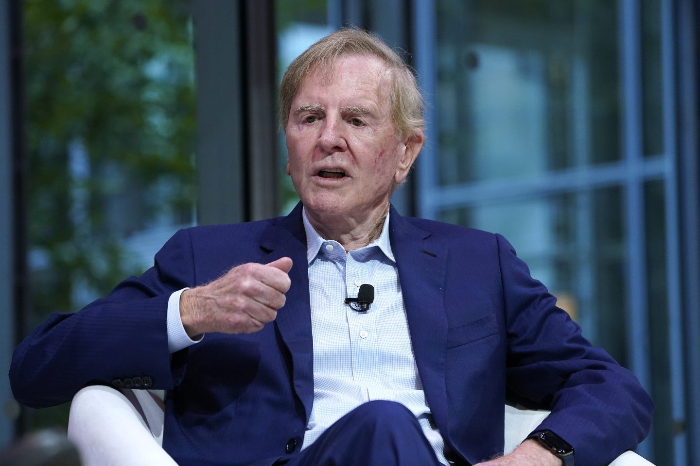 John Sculley, wearing a suit and sitting in a white chair, gestures and speaks with a furrowed brow.