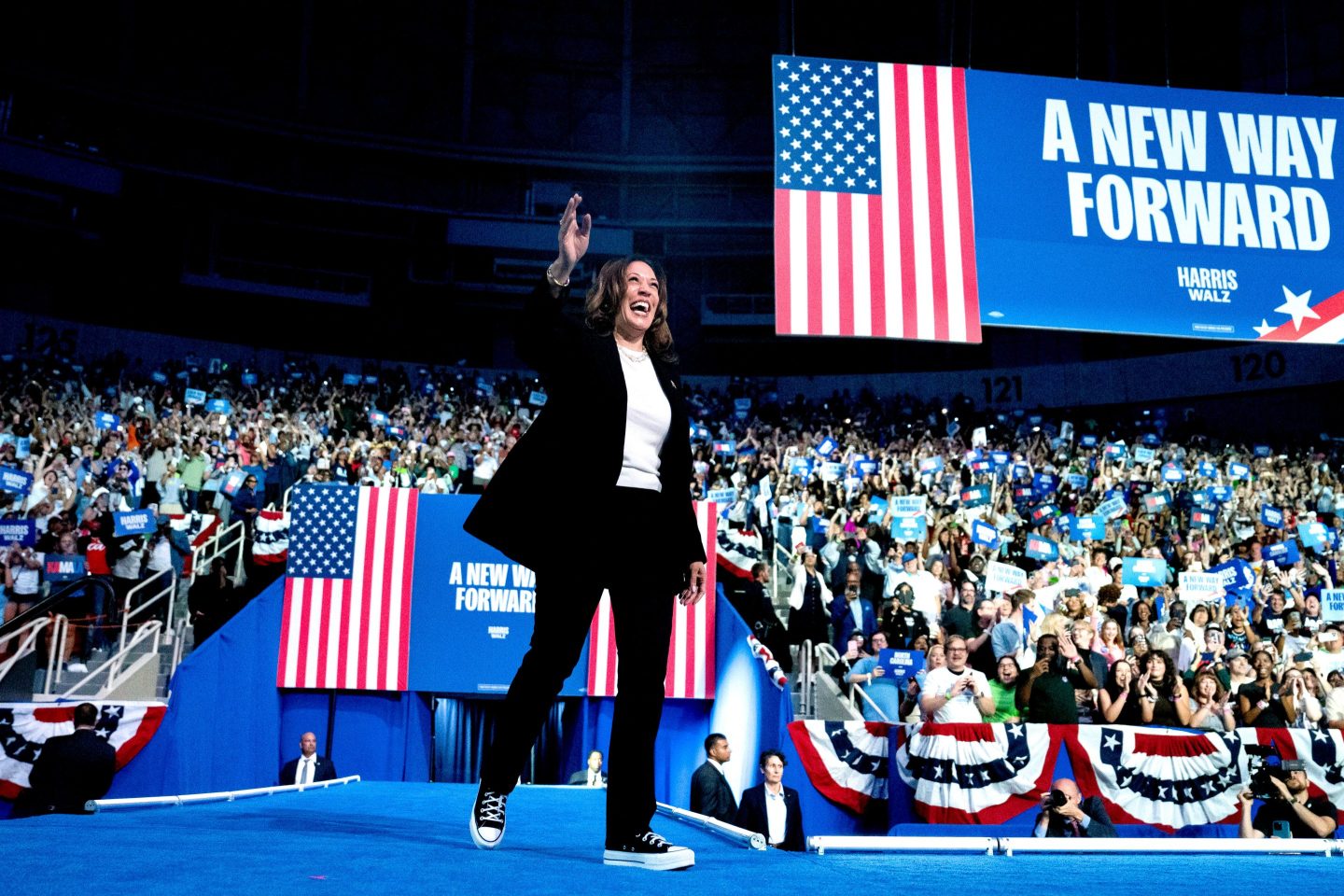 A woman walks across an event stage waving at audience members with a podium and multiple American flags behind her.