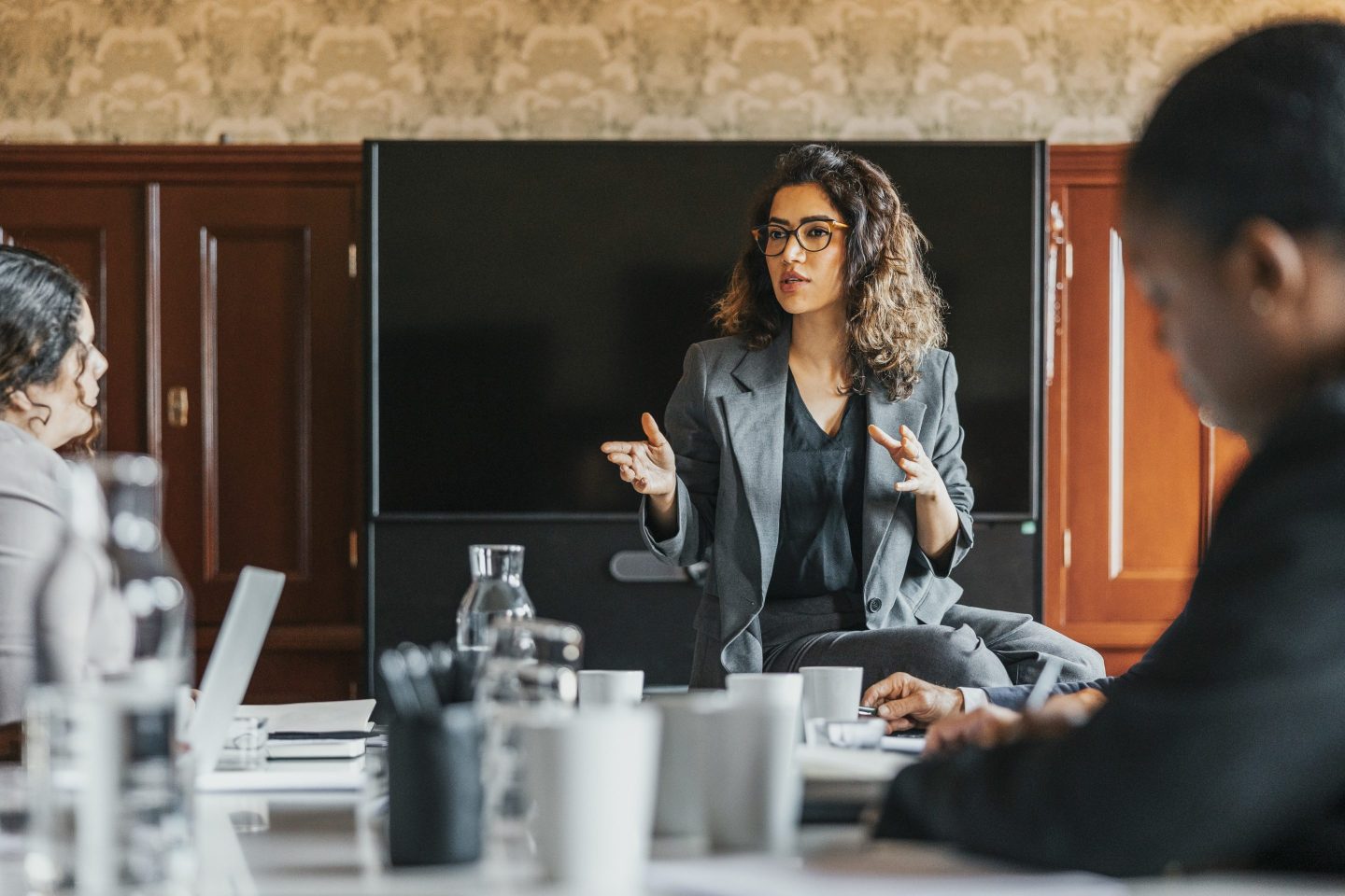 A woman in a dark blazer speaks in front of a table of people.