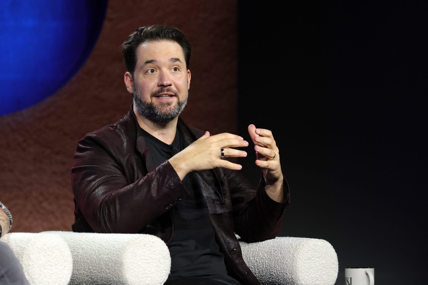 Alexis Ohanian sits in a white chair on stage and gestures with both hands as he speaks.