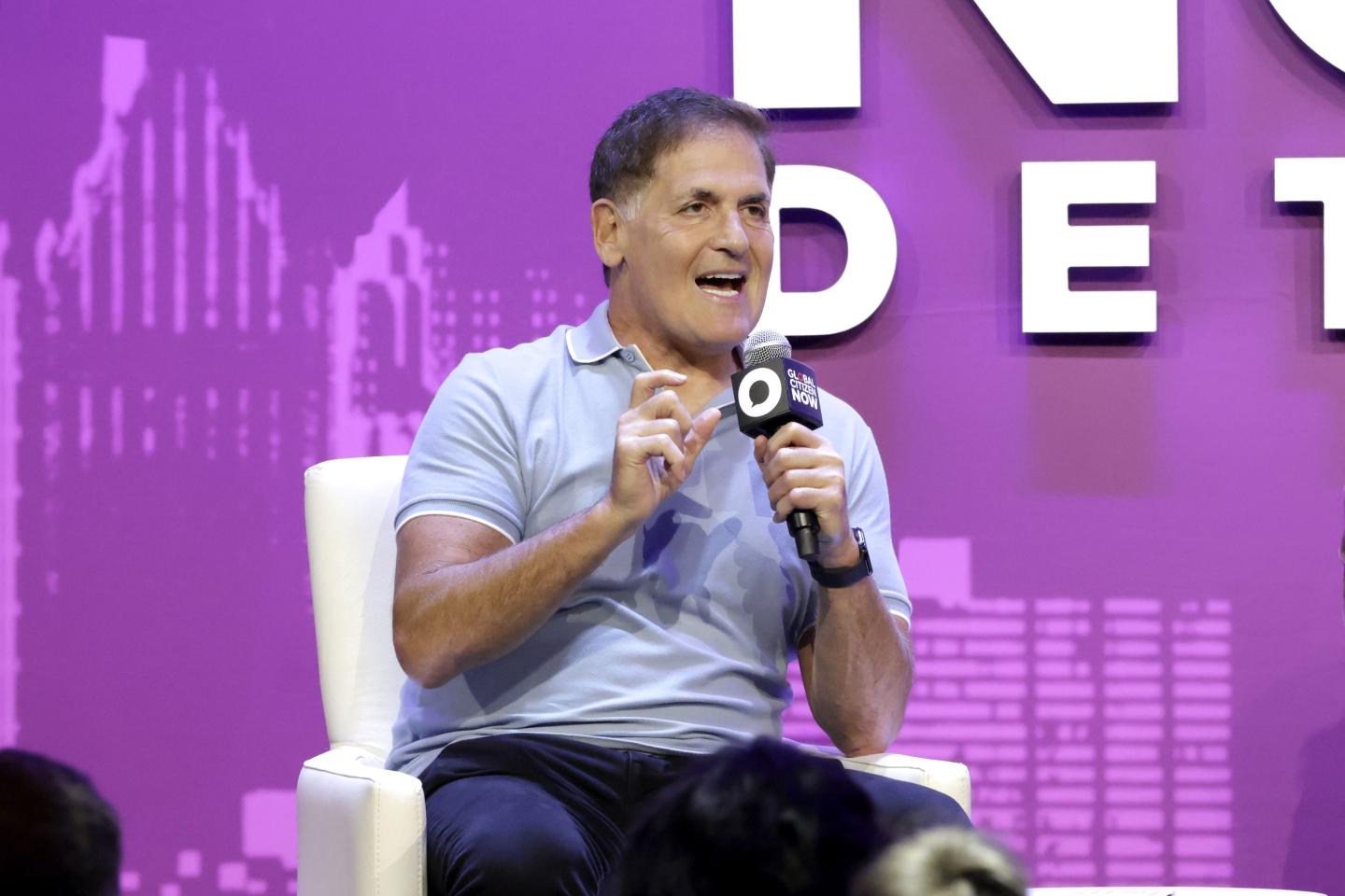 Mark Cuban sits on a white chair in front of a purple background, speaking into a microphone.