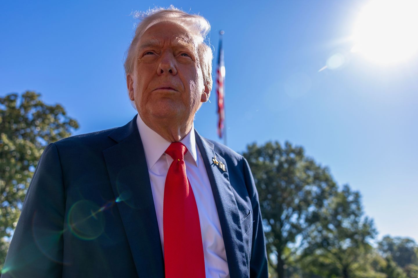 Trump in a suit and red tie with an American flag behind him and a backdrop of trees