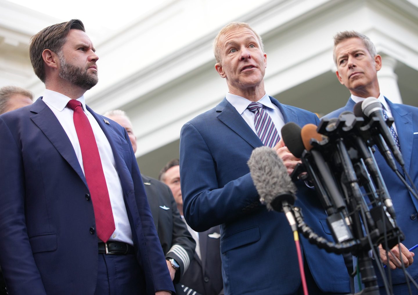 JD Vance and Sean Duffy stand on either side of Scott Kirby as he speaks into a cluster of microphones.