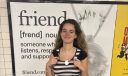 Eva Roytburg shows off her friend.com necklace in front of a poster advertising the friend