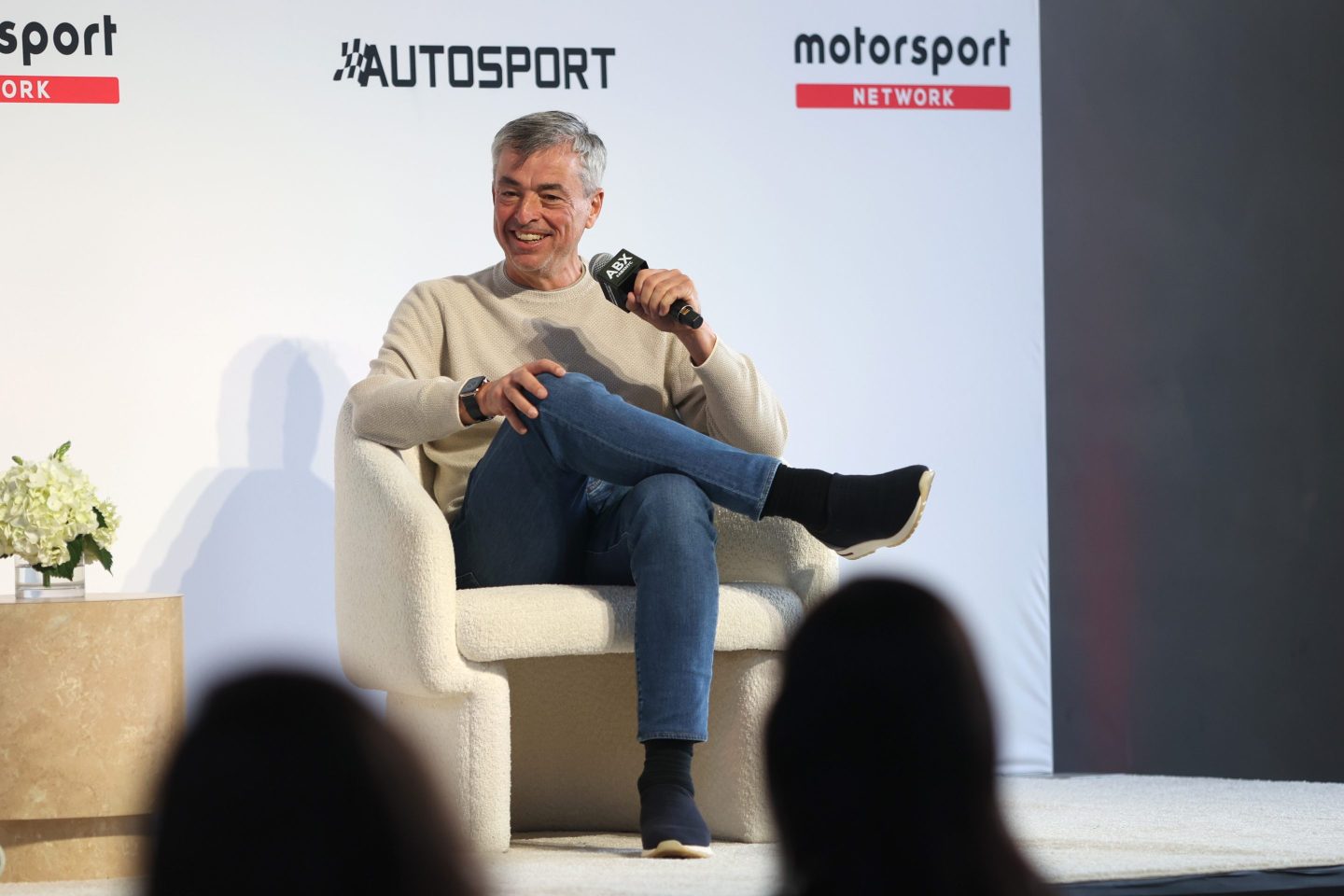 Eddy Cue sits in a white chair on stage, smiling and holding a microphone to his face.
