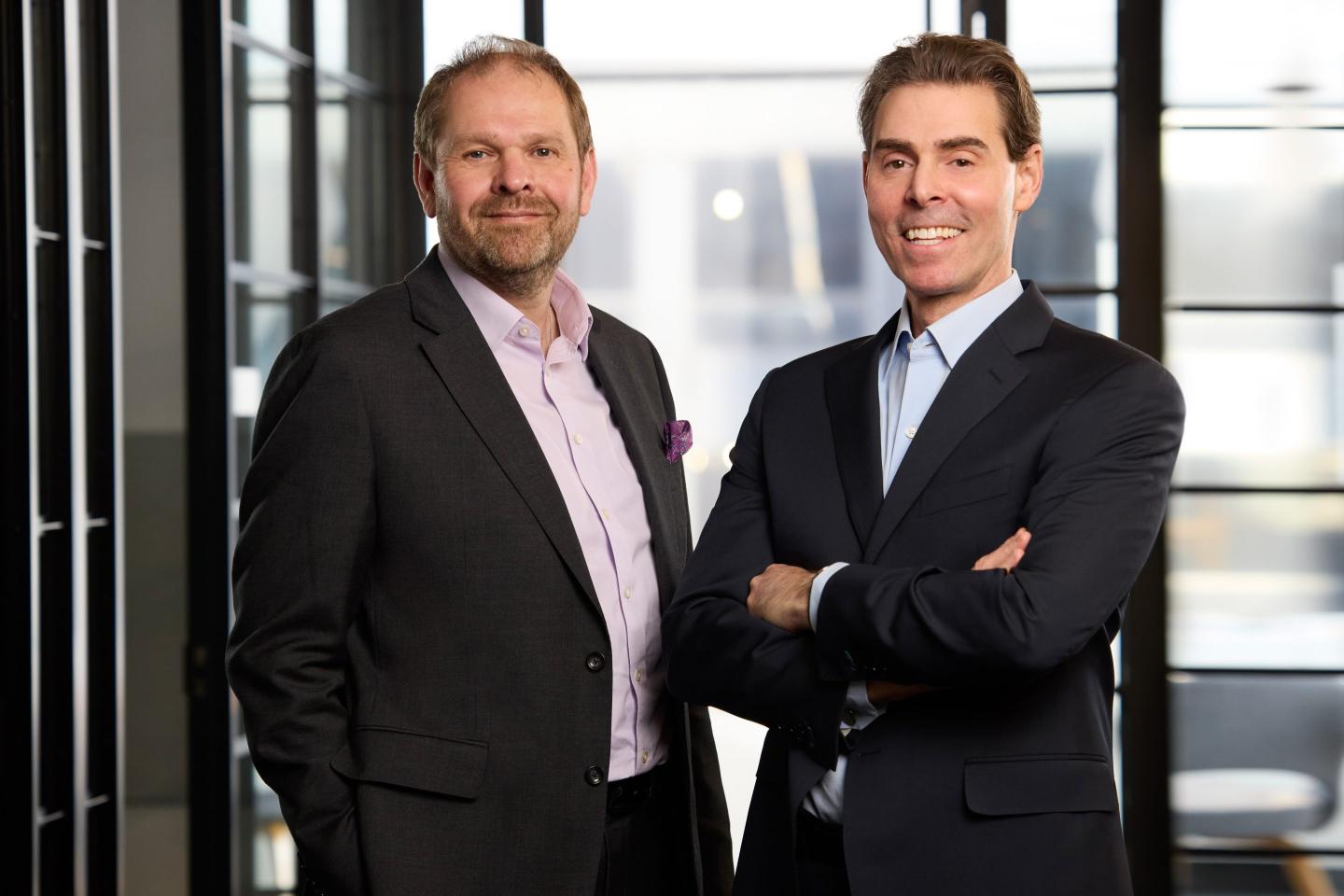 two men in dark suits standing in front of a window