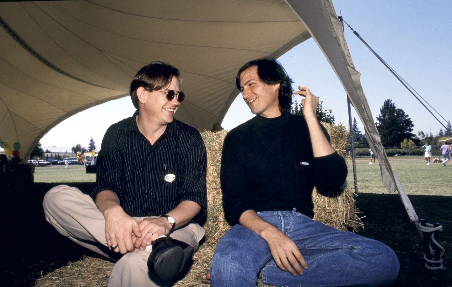 Two men sit facing each other on a hay bale under a tent.