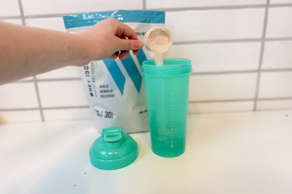A green plastic cup sitting on a counter. A hand is reaching out and pouring Swolverine Whey Isolate powder into the cup with a scooper.