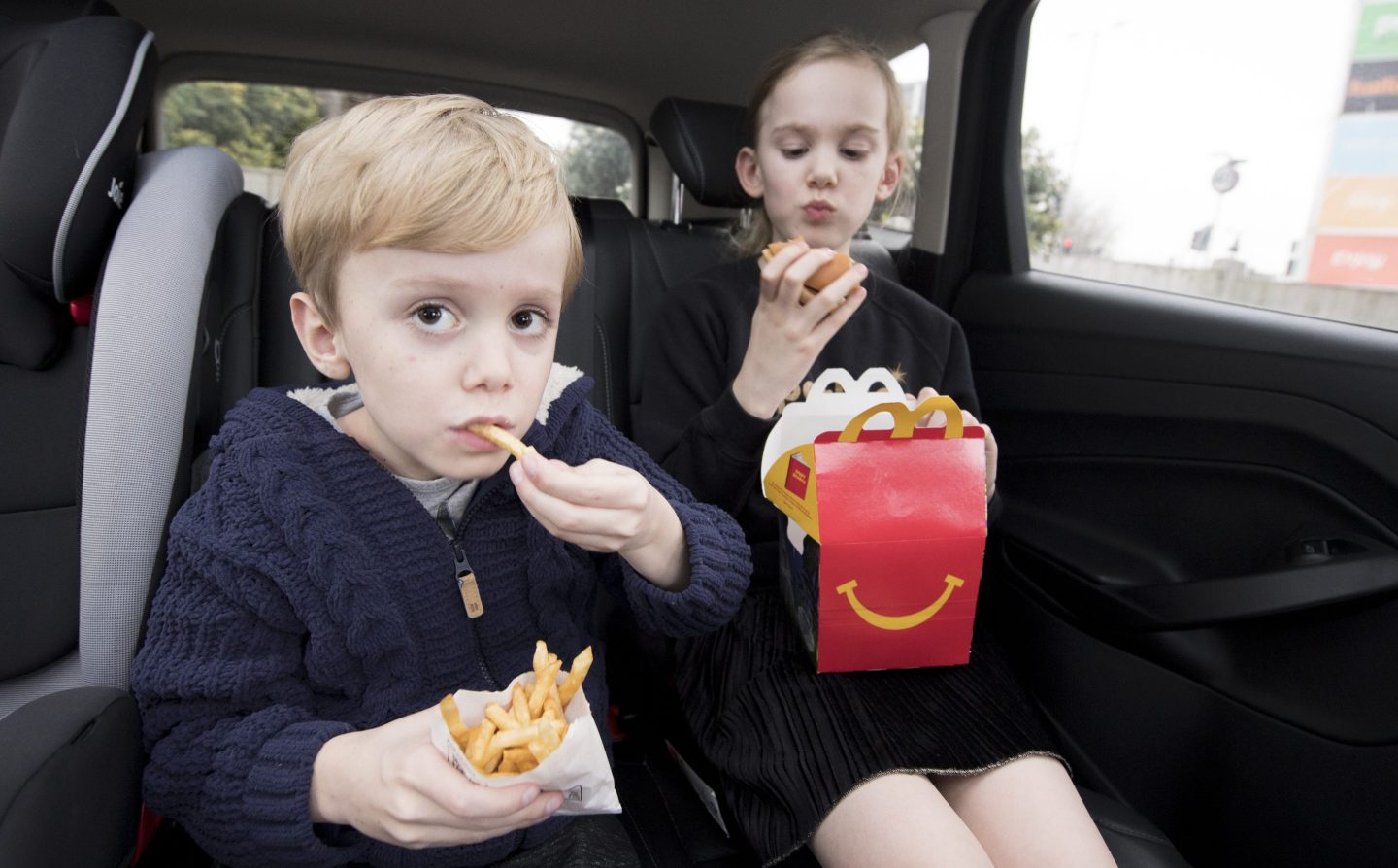 Two kids sit in the back of a car, eating McDonald's.