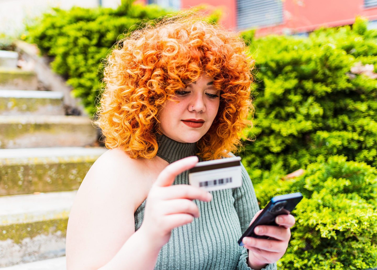 A woman with red, curly hair stands outside and types her credit card information into her phone.