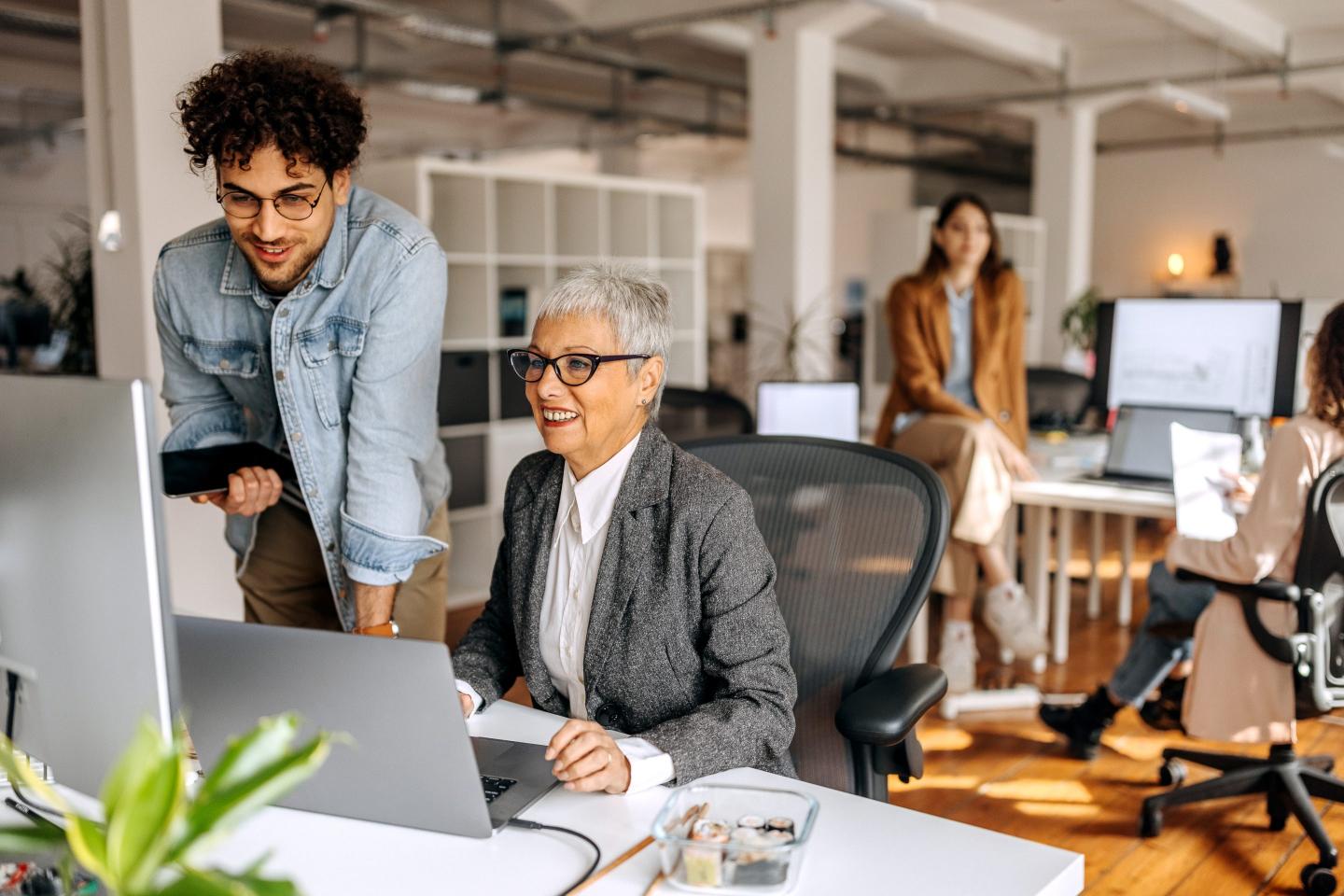 A younger worker helps an older employee at her desk.