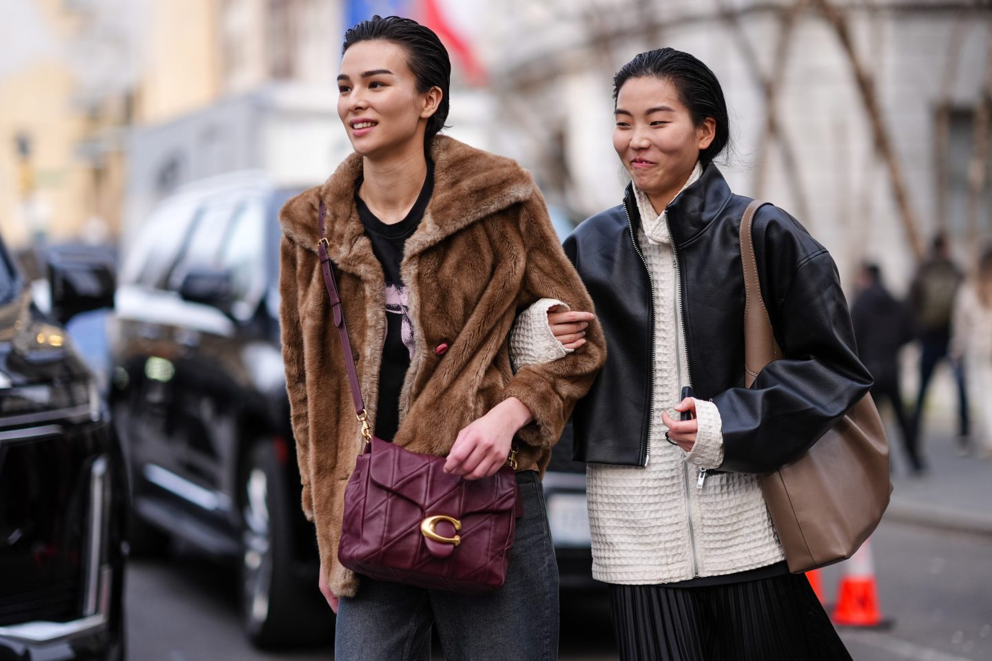NEW YORK, NEW YORK - FEBRUARY 12: A model (L) wears a brown fluffy faux fur coat a black t-shirt, a burgundy / purple leather Coach bag, gray denim jeans pants ; a model (R) wears a black leather jacket , a white knitted / texture jacket , a black pleated skirt , outside Coach, during New York Fashion Week, on February 12, 2024 in New York City.
