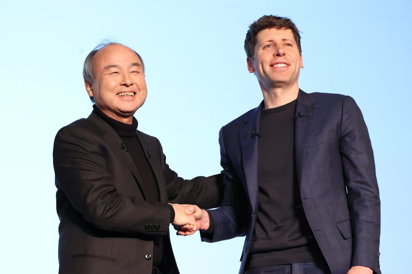 Masayoshi Son and Sam Altman shake hands and smile for the cameras on stage