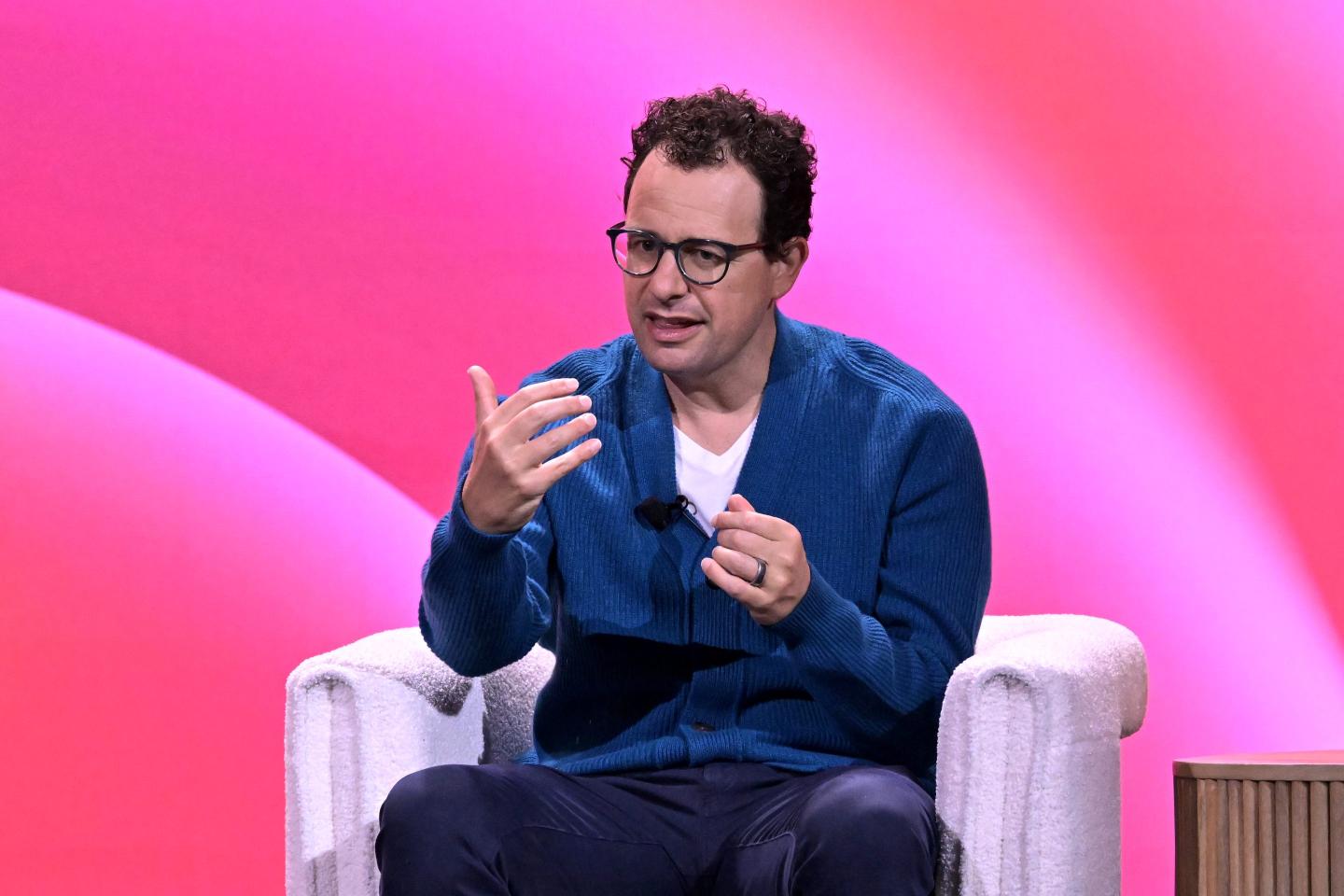 Dario Amodei sits in a white chair in front of a pink background and speaks animatedly.