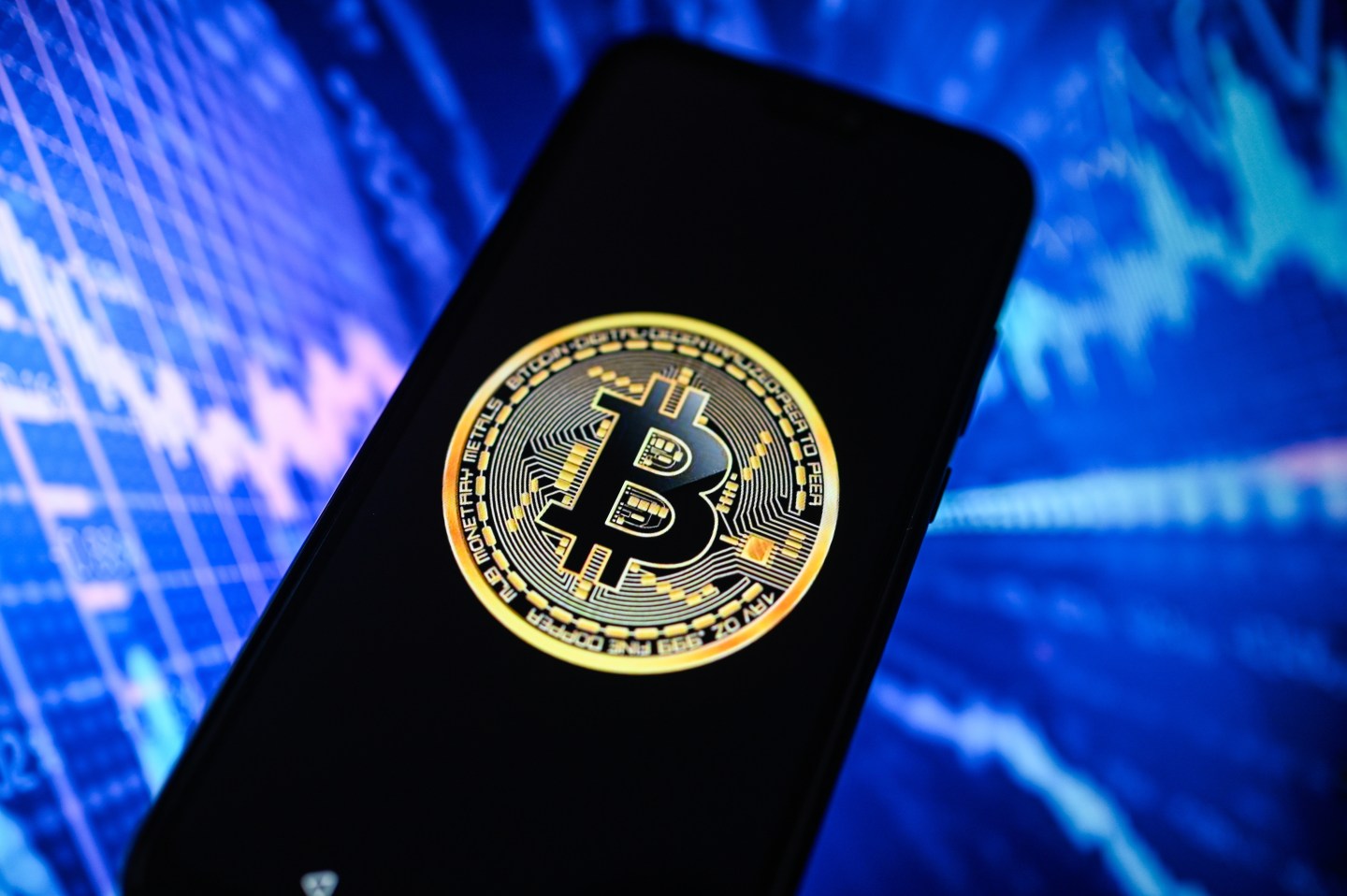 Bitcoin logo is displayed on a smartphone with an illustration of graphs in the background.