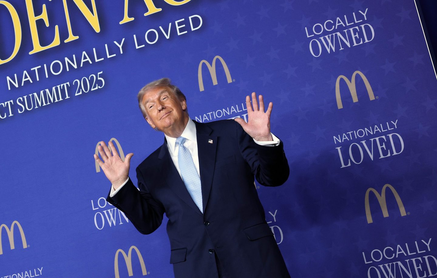 Donald Trump, standing in front of a blue background with the McDonald's logo on it, puts his hands up and smiles.