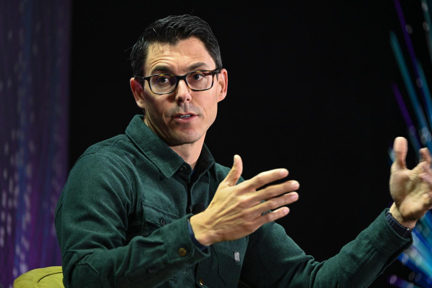 RJ Scaringe, wearing a dark green shirt and glasses, gestures with both hands as he speaks.