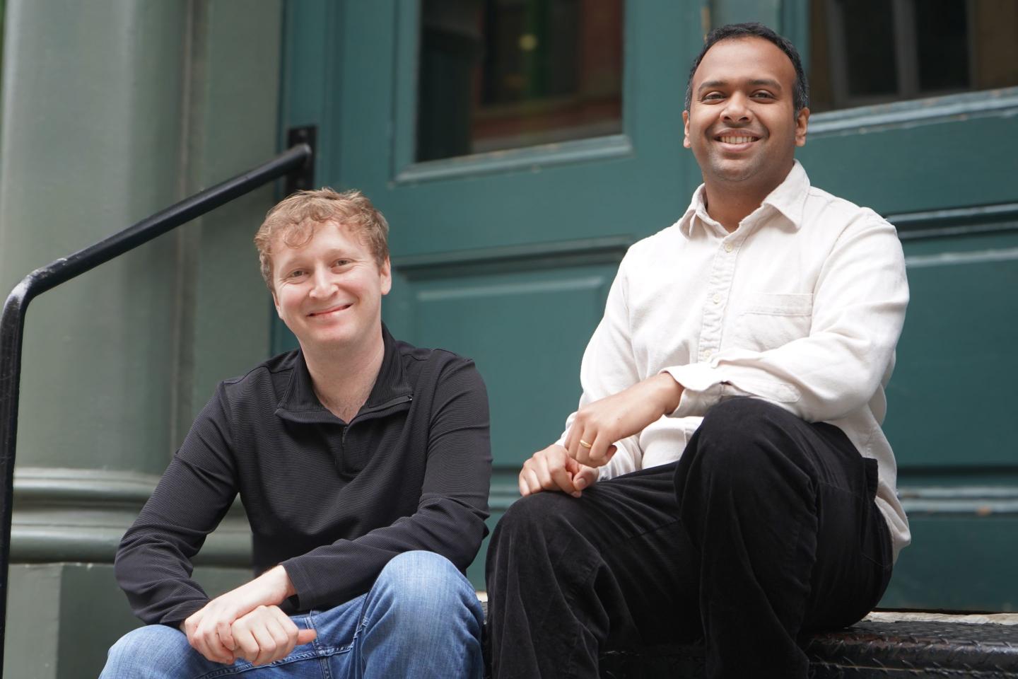 Two men sit and smile in front of a building