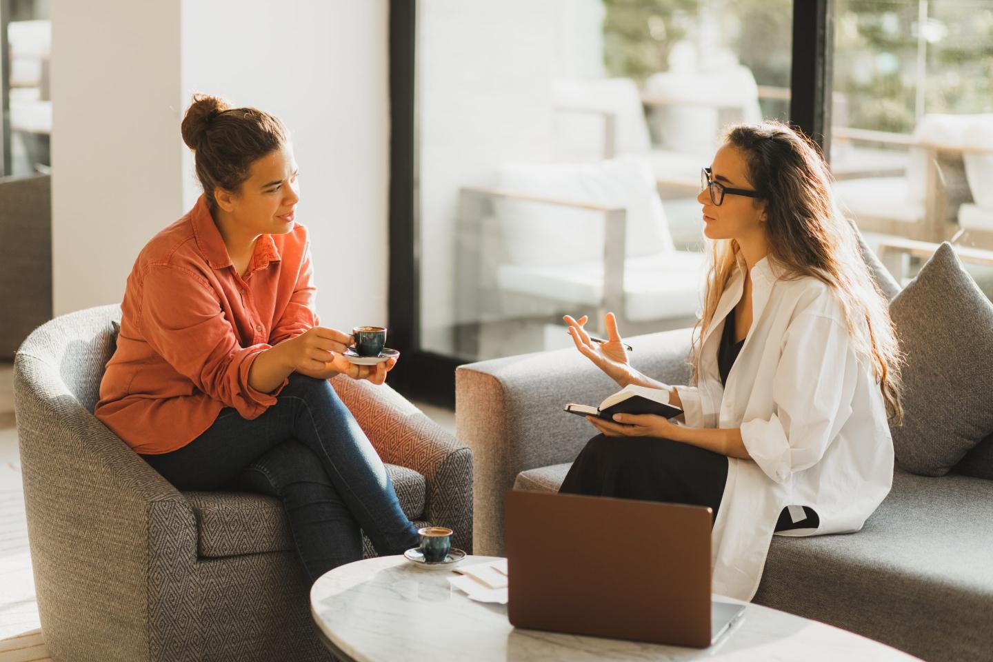 Two women sit in an office talking over coffee.