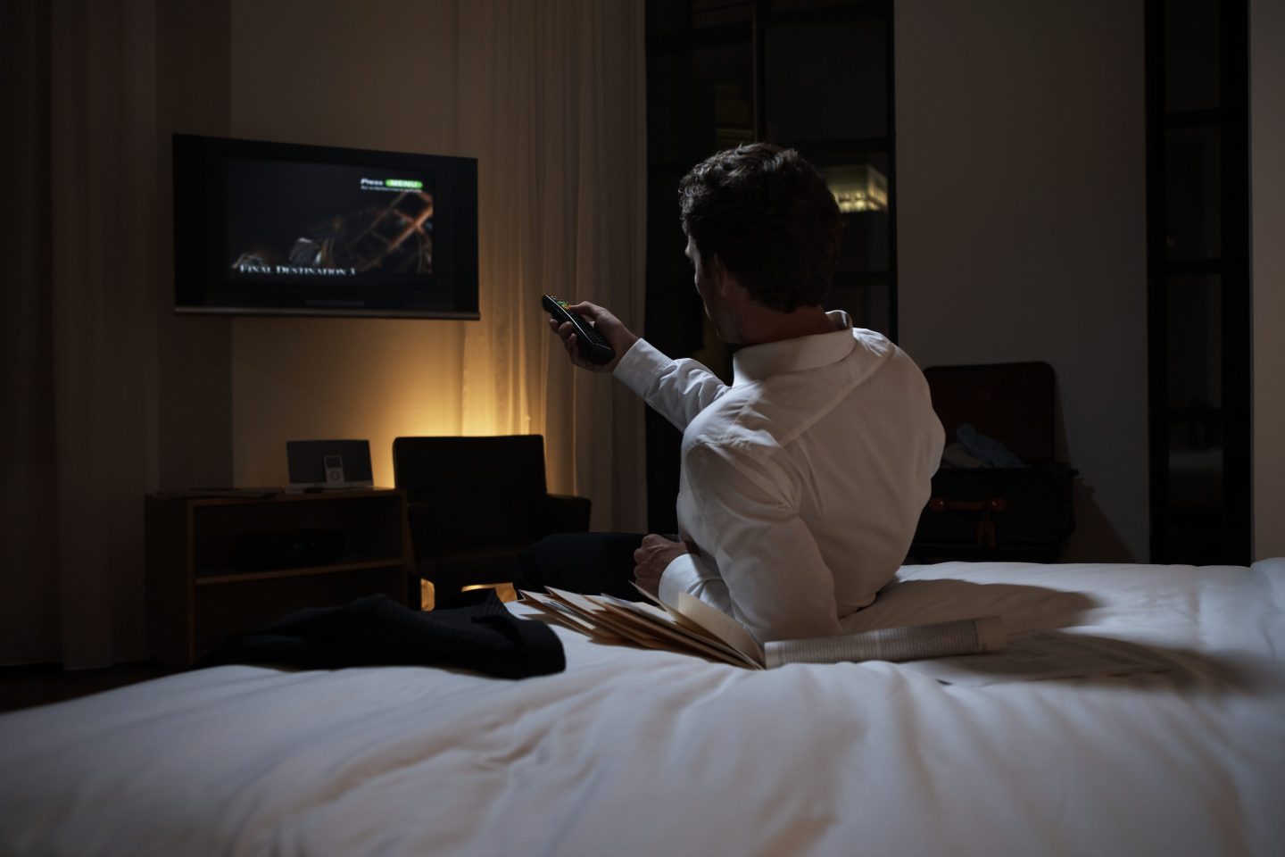 A man on a hotel bed turning on his room's TV