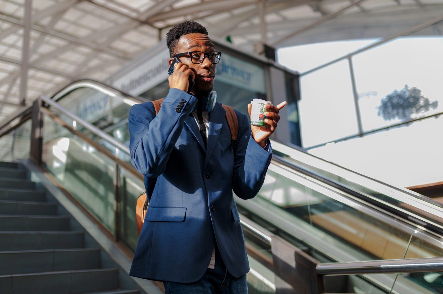 Young businessman walks through a station to catch public transportation to get to his job while talking on the phone and drinking coffee.