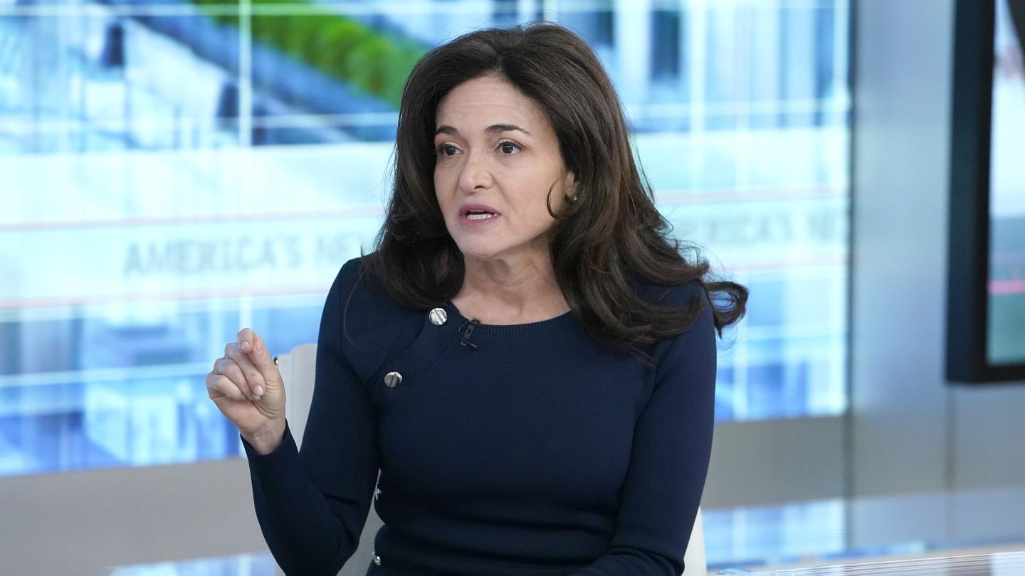 Sheryl Sandberg points with one hand as he sits in front of a light blue background during an interview.