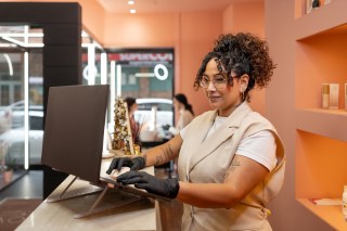 Stylish beautician in black gloves working on a laptop at the front desk of a beauty salon, scheduling appointments