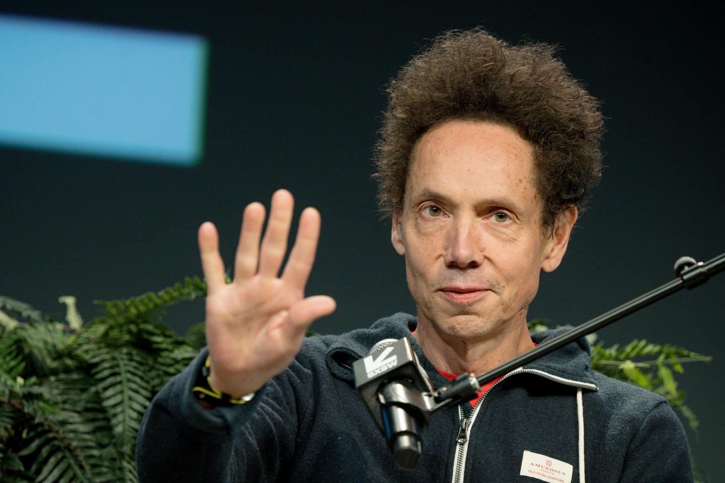 Malcolm Gladwell, sitting behind a microphone, holds his hand up next to him.