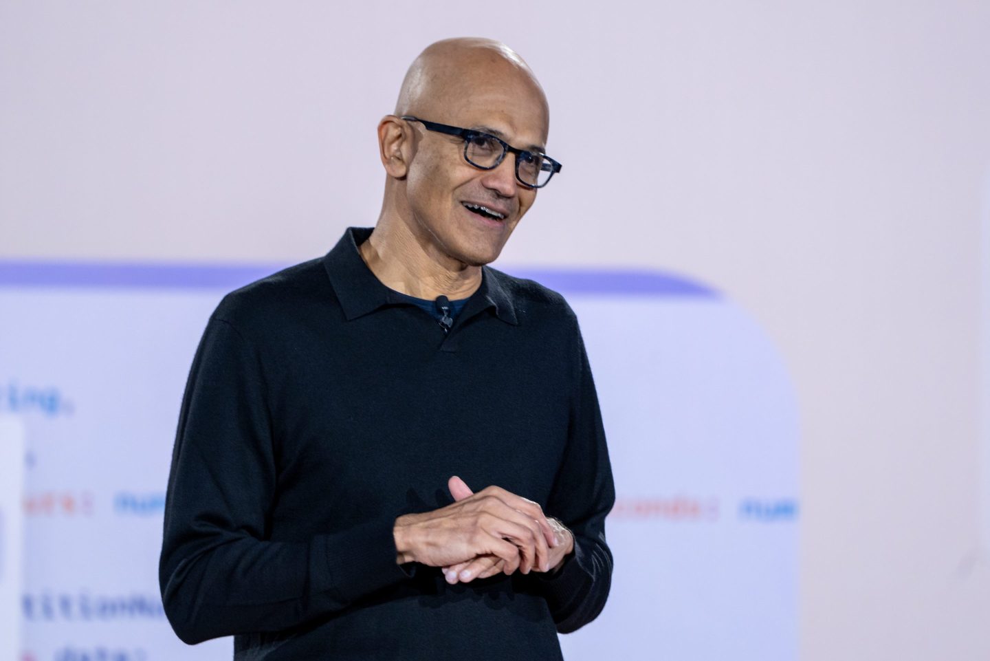 Satya Nadella clasps his hands together and speaks in front of a lilac background on stage.