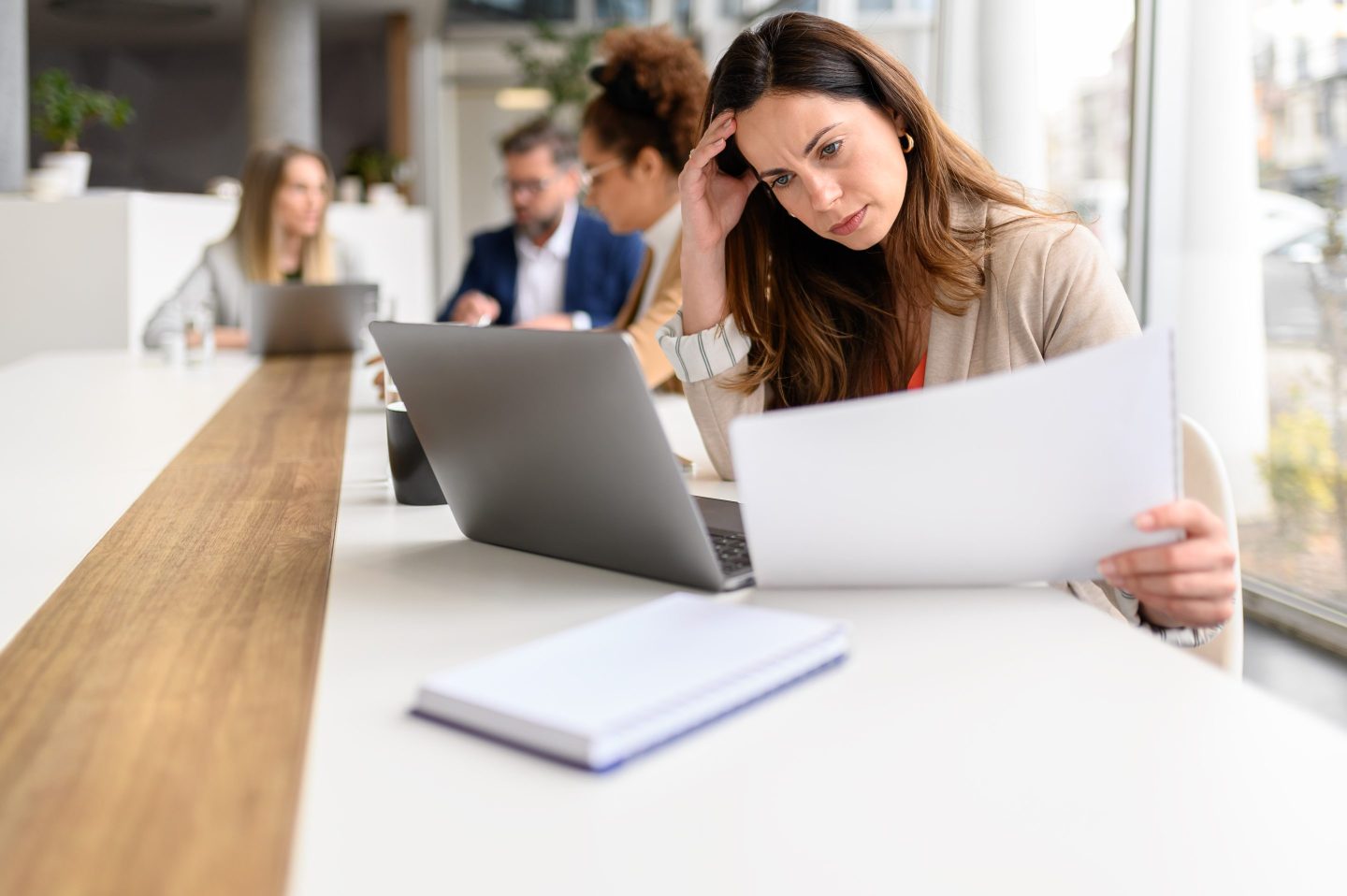 Stressed businesswoman reviewing financial reports while working over laptop in meeting with colleagues in the background