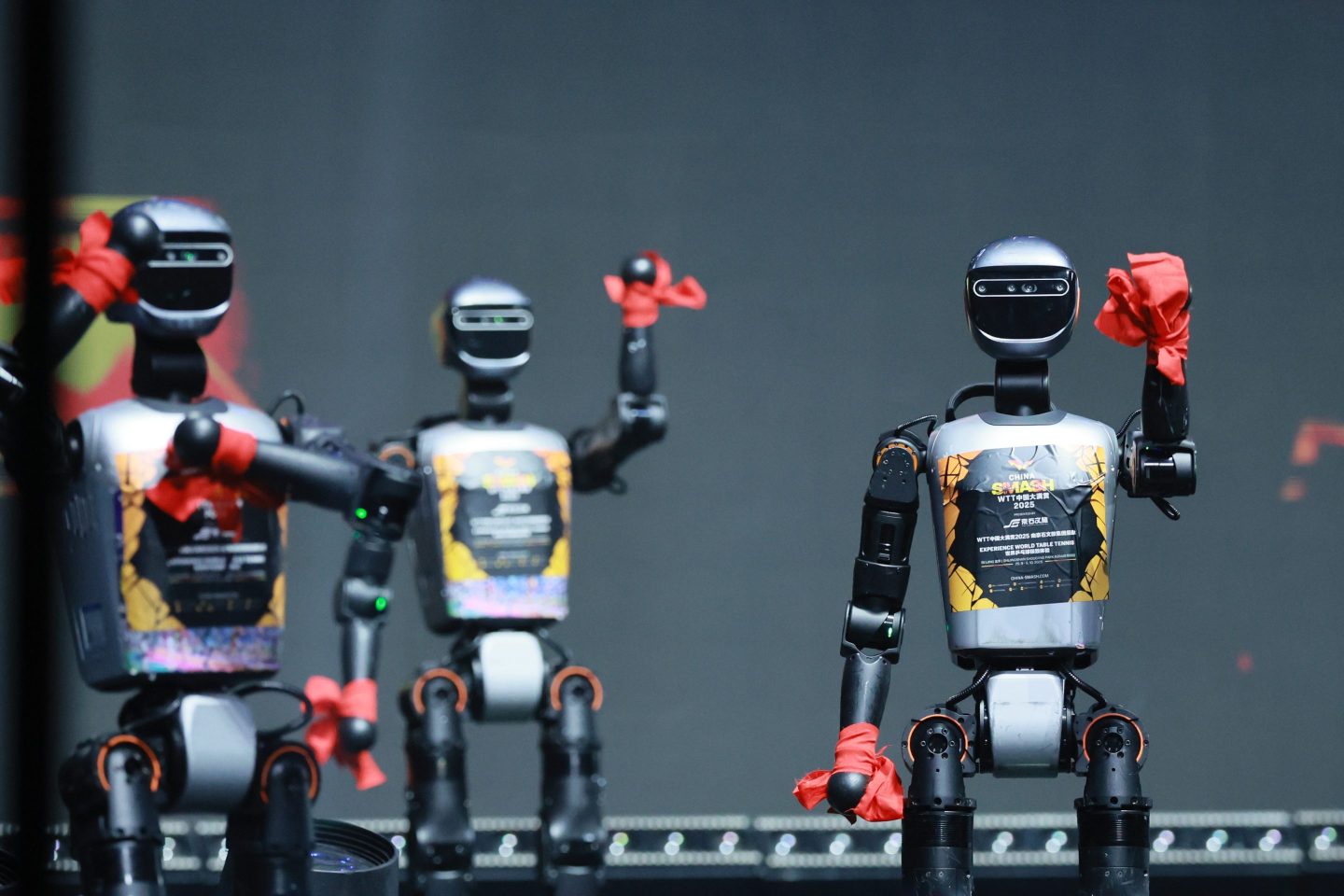 A group of three robots waiving hello to the audience from a stage.