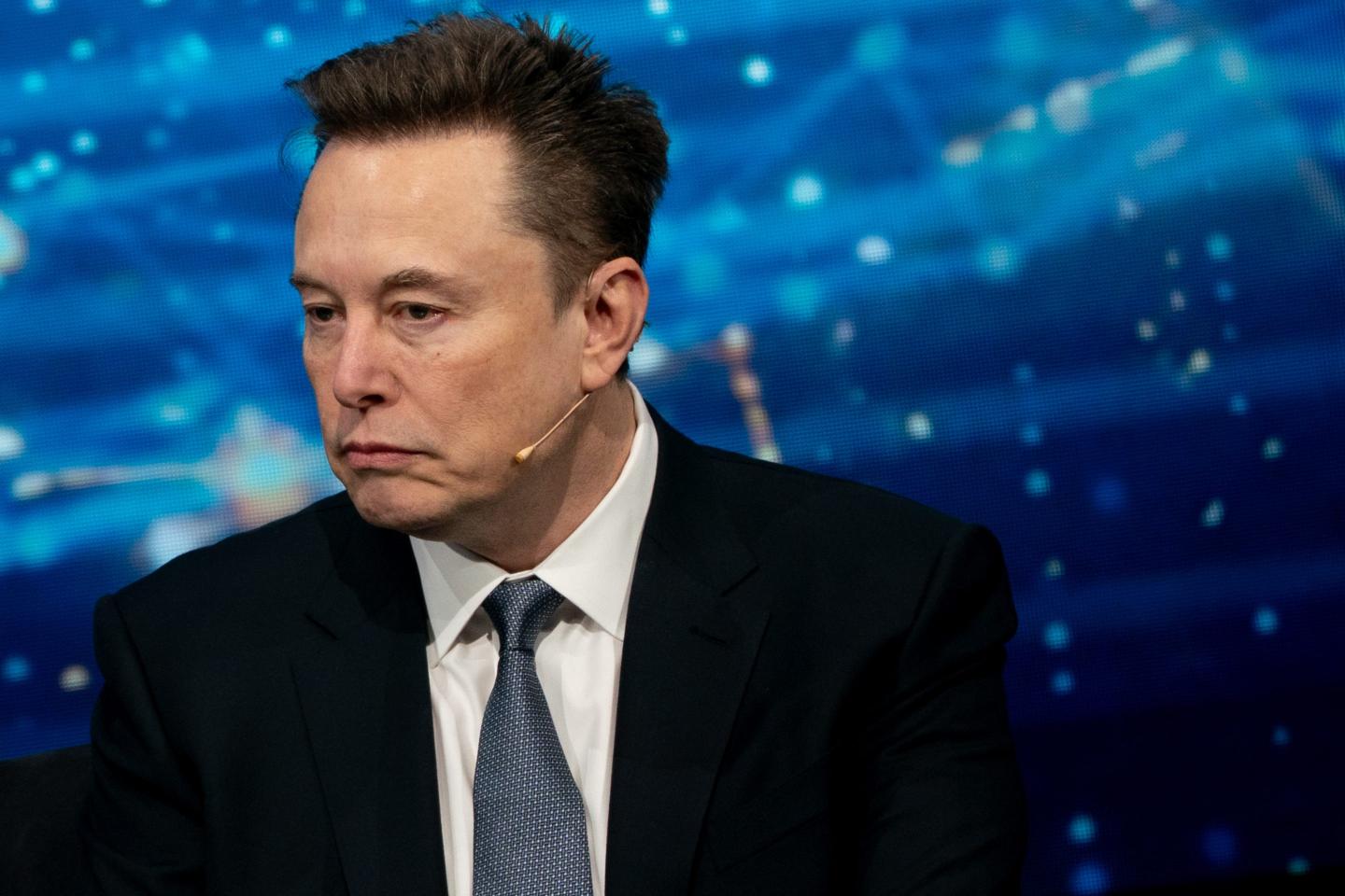 Elon Musk, wearing a suit and in front of a dark blue background, looks to the side and frowns.