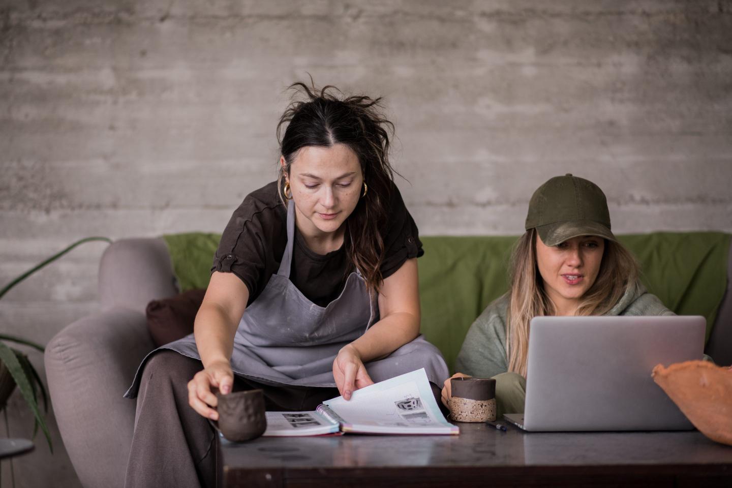 Two female employees, one pointing at a book, other looking at laptop.