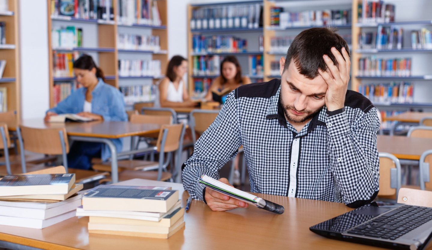 Portrait of upset unhappy diligent  tired male student preparing for exam in university library