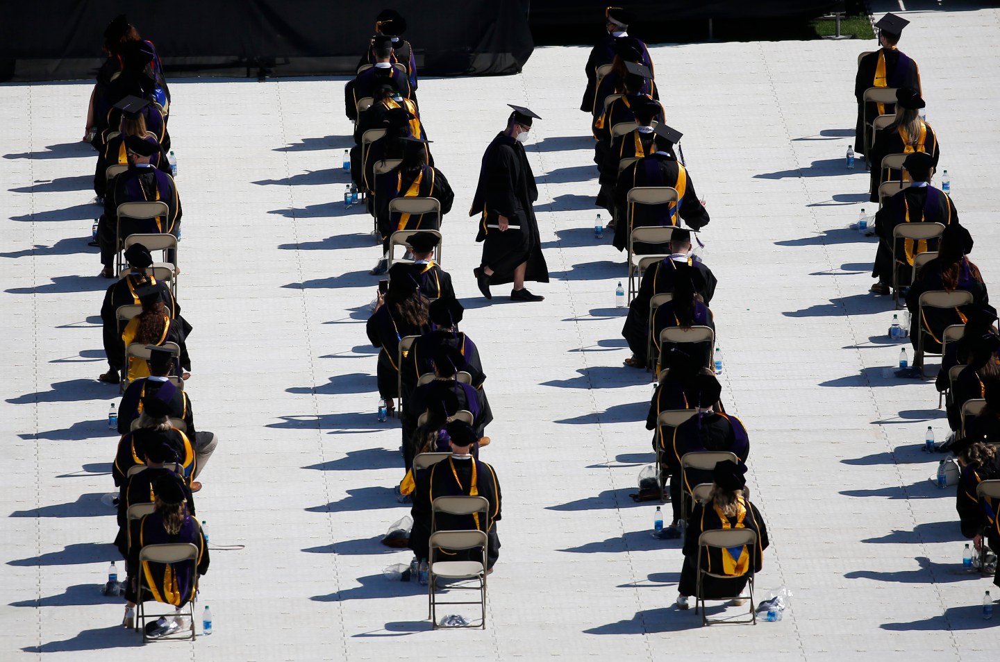 Students sit in lines of chairs in graduation caps and gowns as one student walks across the line of chairs holding a diploma.