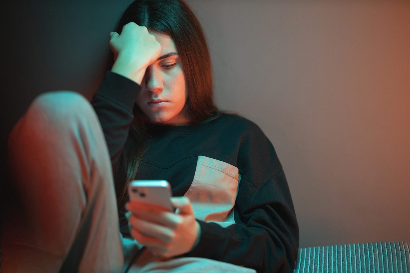 Close-up of a depressed teenage girl sitting on the bed in her room lightened with orange and teal neon lights and scrolling social media.