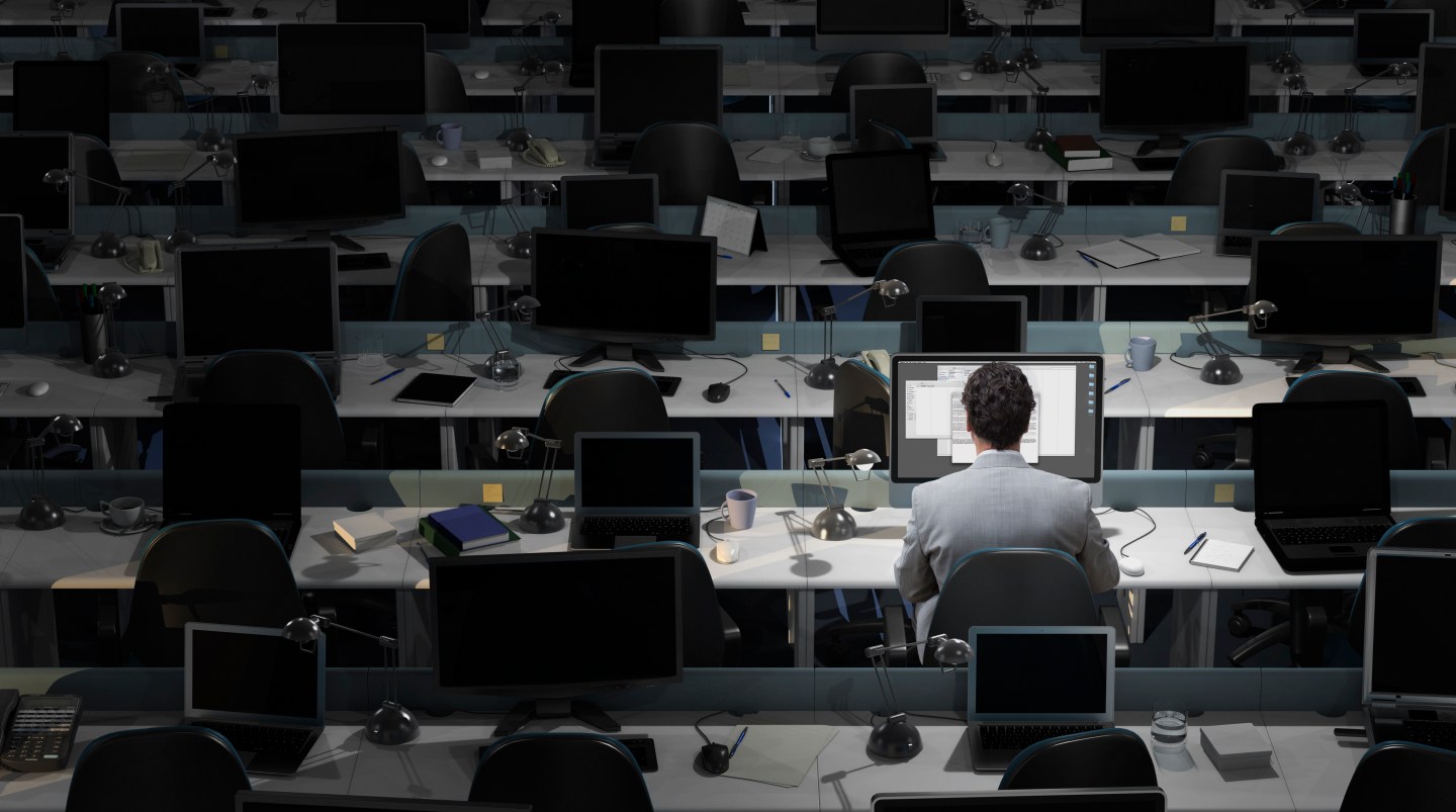 Office worker sits in office surrounded by computers in low lighting. 